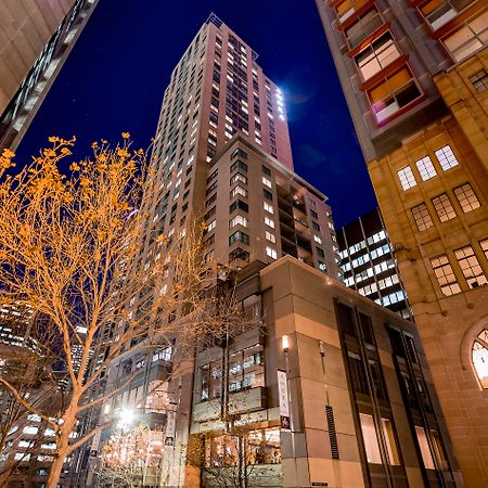 Amora Hotel Jamison Sydney Ngoại thất bức ảnh The photo shows a tall building illuminated at night, surrounded by other structures in an urban environment. The foreground features a tree with bare branches, suggesting it's possibly autumn or winter. The building itself has a modern design, with 