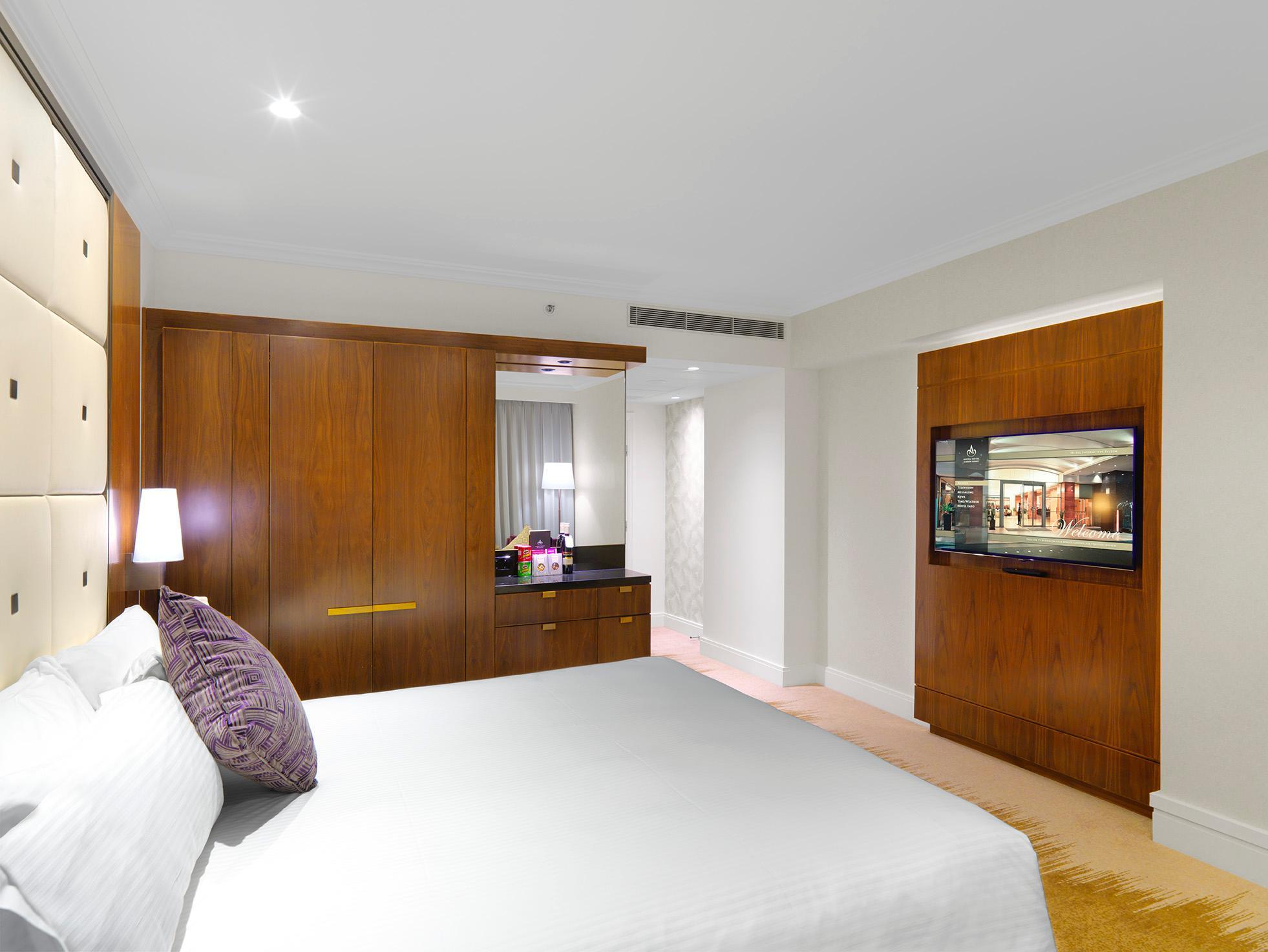 Amora Hotel Jamison Sydney Ngoại thất bức ảnh The photo shows a modern hotel room featuring a neatly made bed with white linens and a decorative purple pillow. The room has wooden finishes, including a built-in wardrobe and a chest of drawers. There is a wall-mounted television on one side and s
