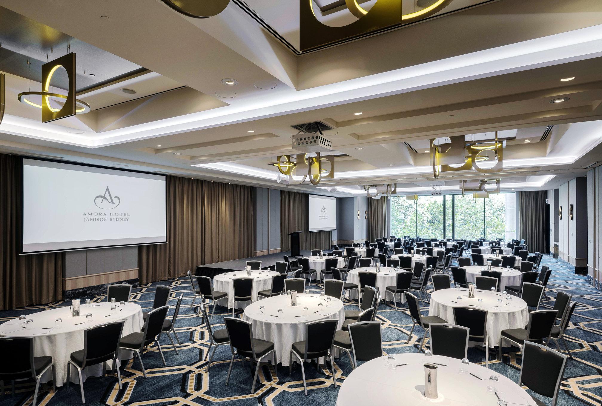 Amora Hotel Jamison Sydney Ngoại thất bức ảnh The photo shows a spacious conference or event room. There are multiple round tables arranged with white tablecloths and black chairs around them. The room has a modern design with elegant lighting fixtures hanging from the ceiling. A large screen is