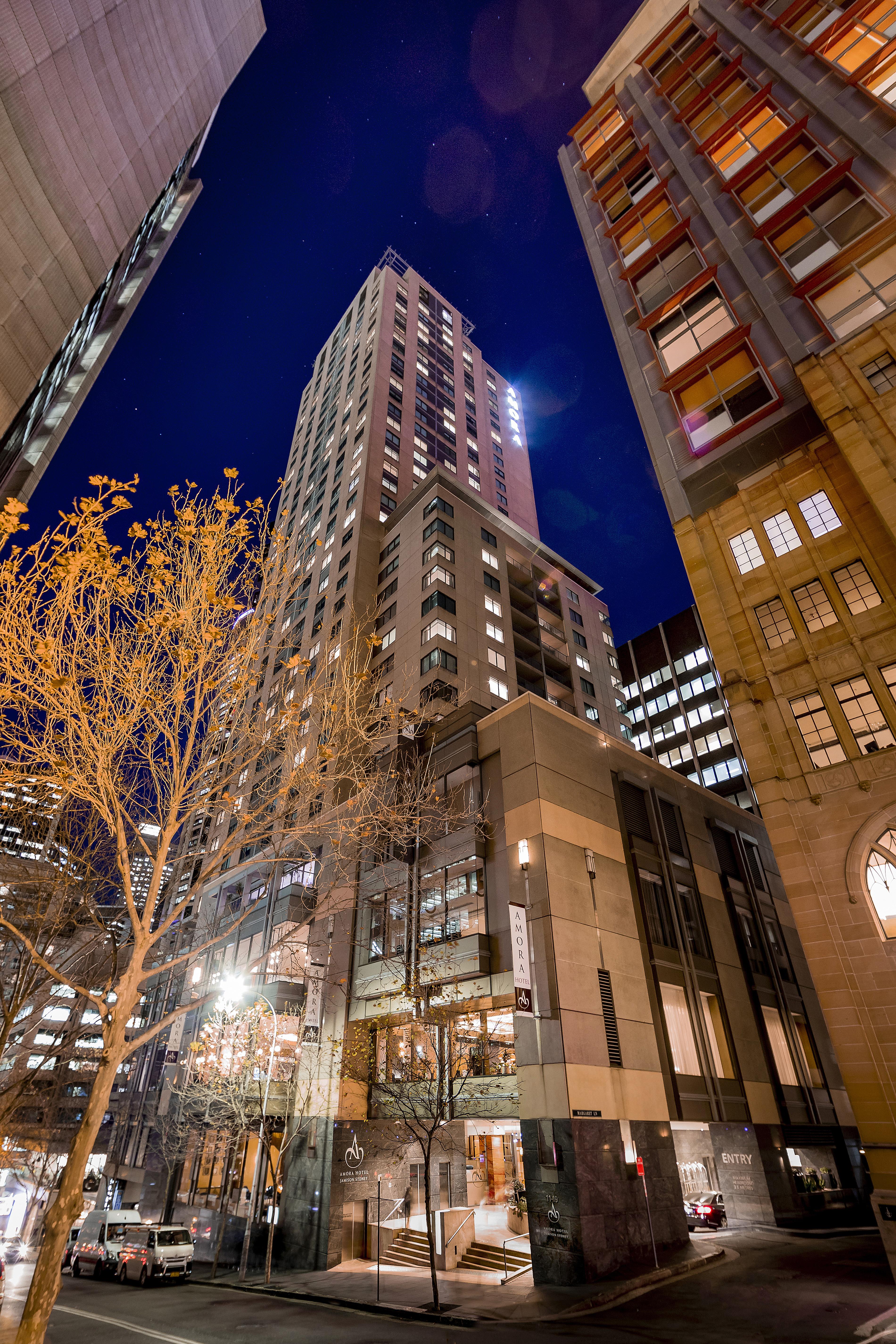 Amora Hotel Jamison Sydney Ngoại thất bức ảnh The photo shows a tall building illuminated at night, surrounded by other structures in an urban environment. The foreground features a tree with bare branches, suggesting it's possibly autumn or winter. The building itself has a modern design, with 