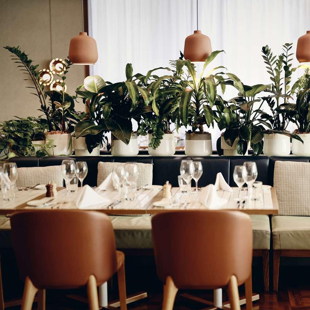 Amora Hotel Jamison Sydney Nhà hàng bức ảnh The photo shows a stylish restaurant interior featuring a warm and inviting atmosphere. There are neatly arranged tables with wooden surfaces, each set with glasses, napkins, and seasonings. Surrounding the tables are large, green plants in white pot