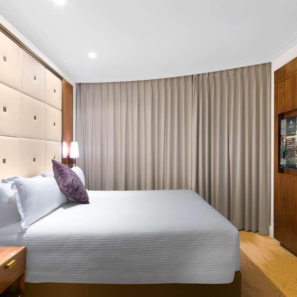 Amora Hotel Jamison Sydney Phòng bức ảnh The photo shows a modern hotel room with a neatly made bed featuring light-colored bedding and a decorative pillow. The headboard behind the bed has a soft, upholstered design with square patterns. To the right, there is a wooden cabinet, and on the 