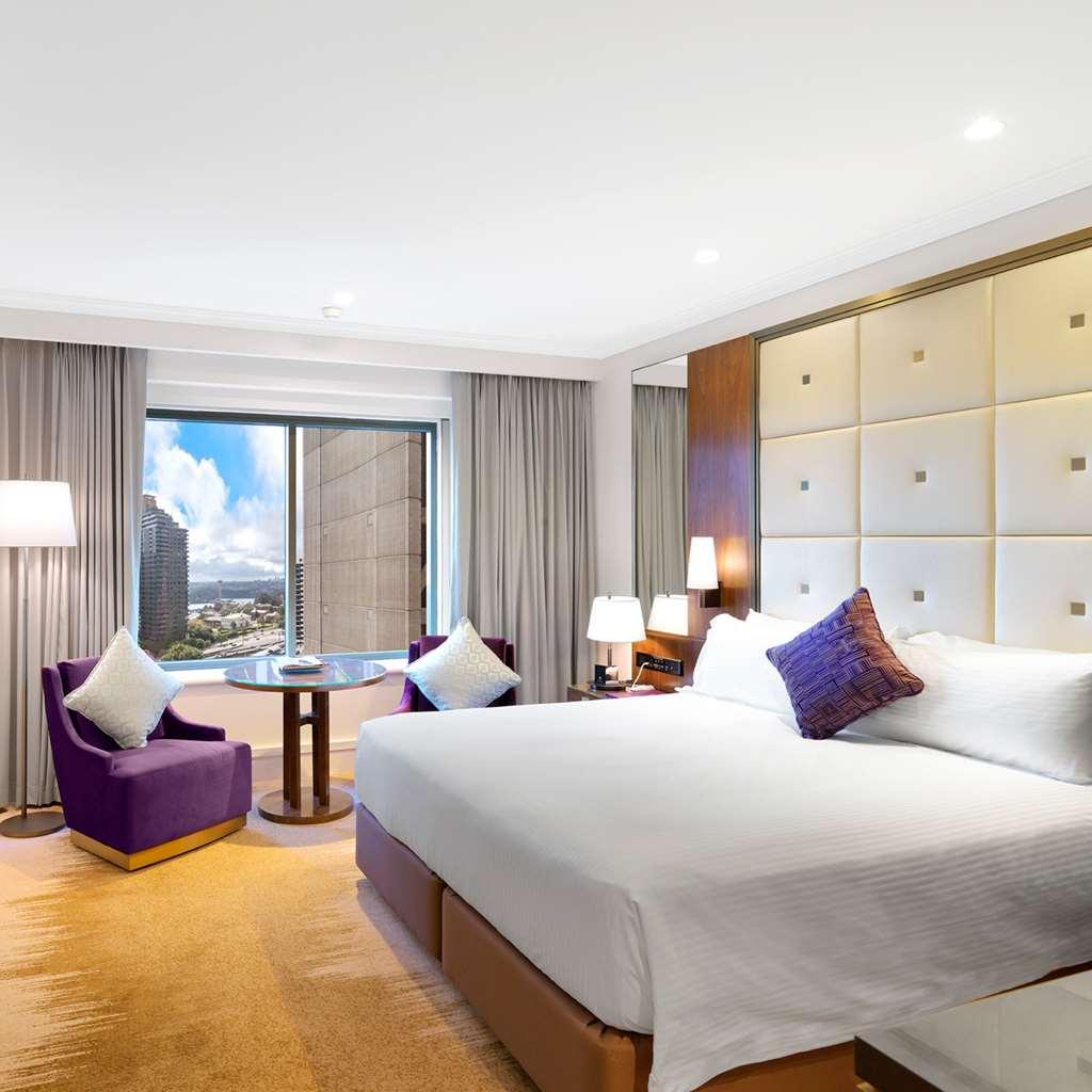 Amora Hotel Jamison Sydney Phòng bức ảnh The photo shows a modern hotel room featuring a king-sized bed with white linens and a decorative purple pillow. To the side, there are two stylish armchairs in a deep purple color, accompanied by a small round table. The room has large windows cover
