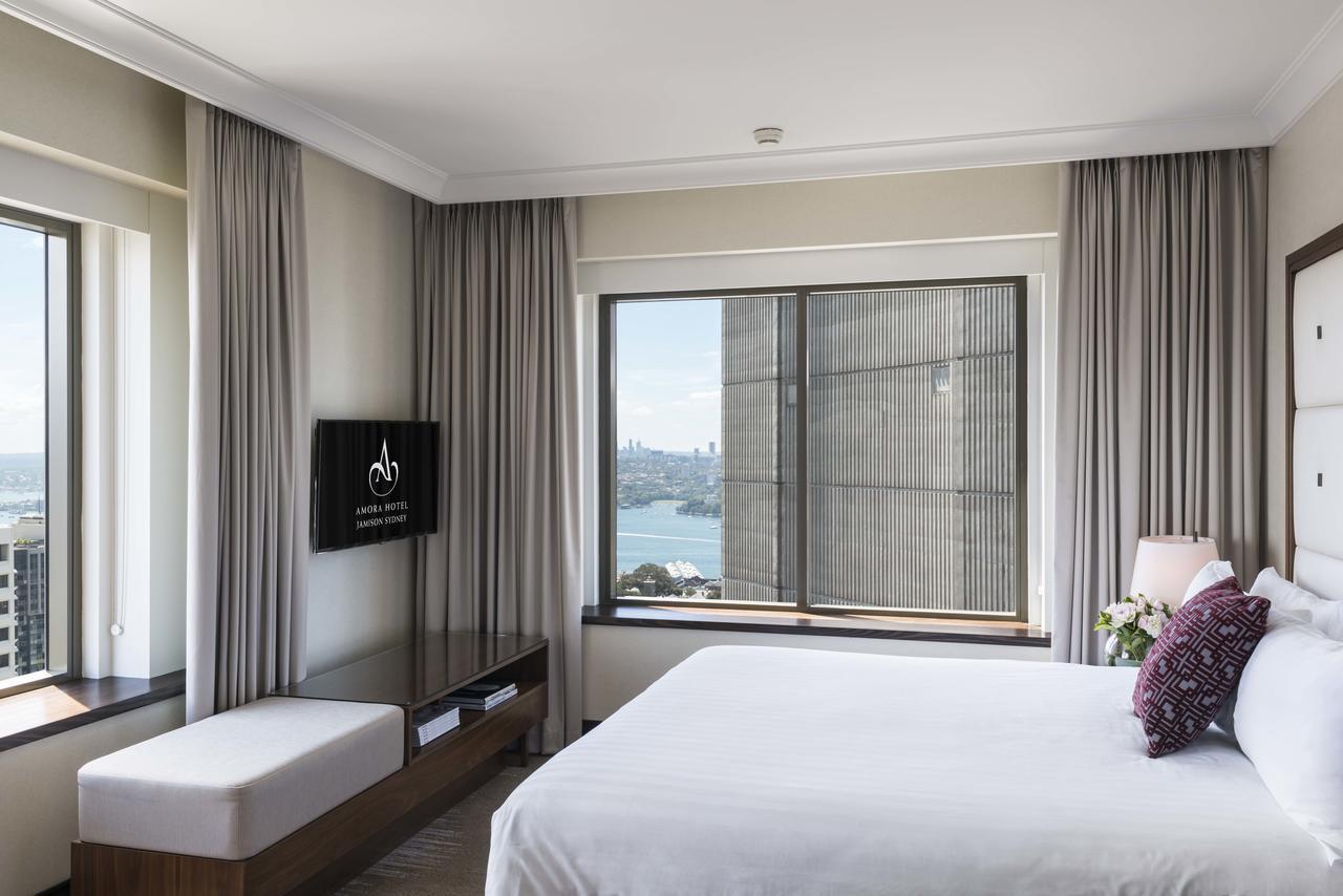 Amora Hotel Jamison Sydney Ngoại thất bức ảnh The photo shows a modern hotel room featuring a large bed with white bedding. There is a seating area with a bench next to a window. The room is well-lit with natural light coming in through the window, which has curtains. A flat-screen TV is mounted