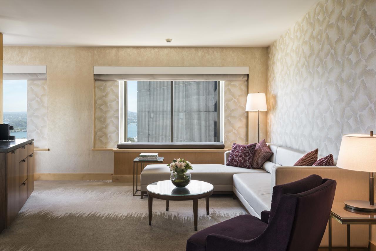 Amora Hotel Jamison Sydney Ngoại thất bức ảnh The photo shows a modern and stylish hotel room or living space. It features a large window with city views and light-colored walls adorned with a subtle pattern. There is a comfortable white sofa accompanied by purple accent pillows, positioned near