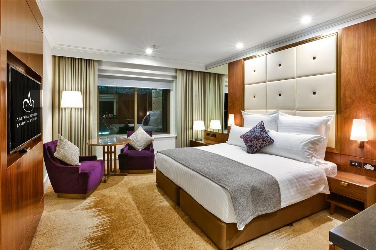 Amora Hotel Jamison Sydney Ngoại thất bức ảnh The photo shows a modern hotel room with a clean and stylish design. It features a large bed with white linens and a gray throw blanket. There are decorative pillows on the bed. To one side, there is a small seating area with two purple chairs and a 