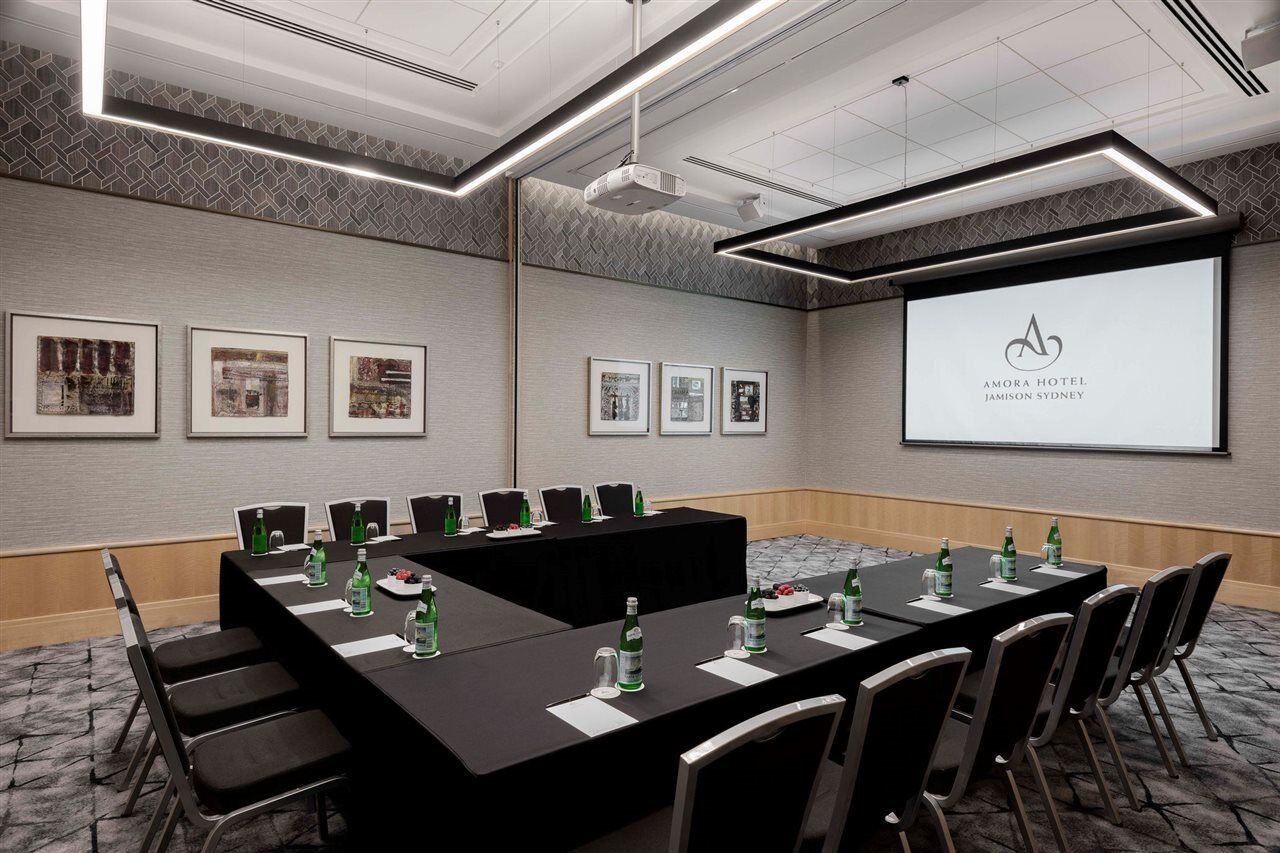 Amora Hotel Jamison Sydney Ngoại thất bức ảnh The image shows a modern conference room setup. 

In the center, there is a large black rectangular table surrounded by chairs, indicating it's arranged for a meeting. On the table, there are several water bottles, cups, and likely some snacks or not